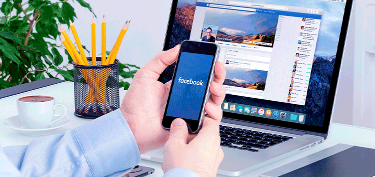 Uma pessoa segurando um celular na frente de um laptop, ambos com o facebok aberto